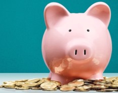 A pink piggy bank on a table in front of a blue wall with gold coins surrounding it.