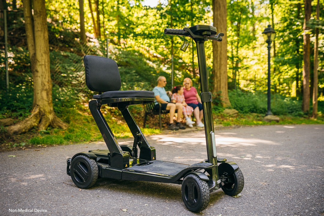 The Go Go Carbon sits outside on a pathway. Behind it, two grandparents and a grandchild sit on a bench in front of trees. 