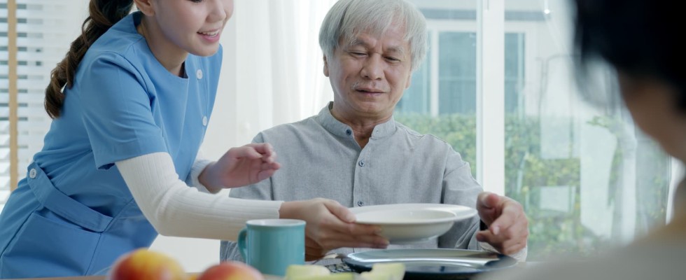 A young woman caregiver is bringing a tray of breakfast food to an older gentleman in his home.