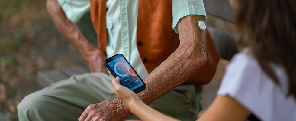 Caregiver helping senior diabetic man check his glucose data on smartphone outdoors in a park.