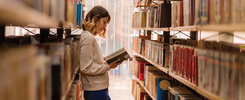 A woman in the stacks at a library, reading a book.