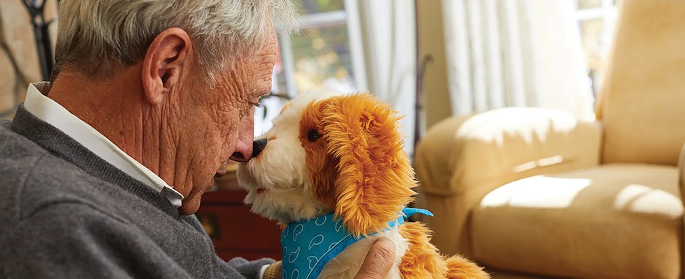 An older man holds a companion pet, which looks like a brown and white spotted dog with a blue bandanna around its neck, up to his face. Touching his nose with the robotic dogs. 