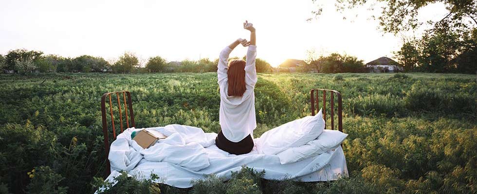 A young woman stretches while sitting on her bed in the middle of an open field.