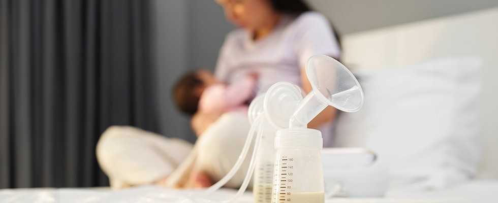 In the foreground, a breast pump is sitting with milk in it. In the background a woman is nursing a baby..