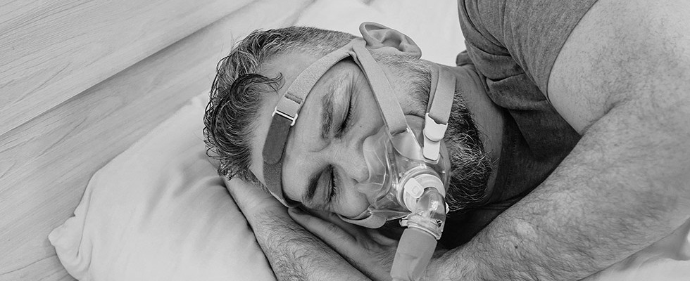 A black and white image of a middle aged man sleeping with a CPAP mask on