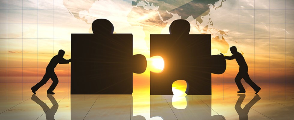 Foreground: Silhouettes of two people pushing together two large puzzle pieces. In the background is a sunset, shining through the gap in the puzzle pieces and a superimposed map of the world. 