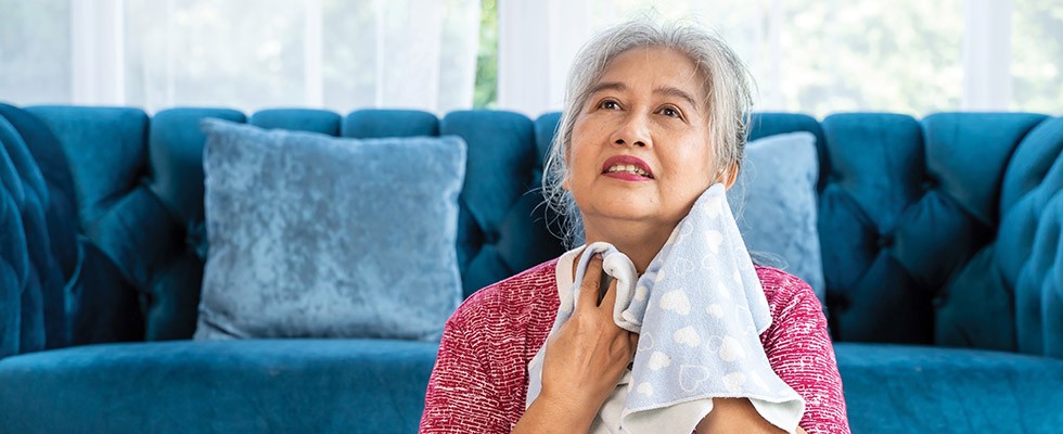 An older woman sits in front of a couch, with a cloth to wipe away sweat.