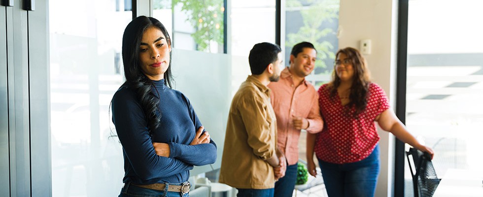 In the foreground, a woman has her arms crossed and looks away, seemingly upset. In the background three people are chatting, close together. 