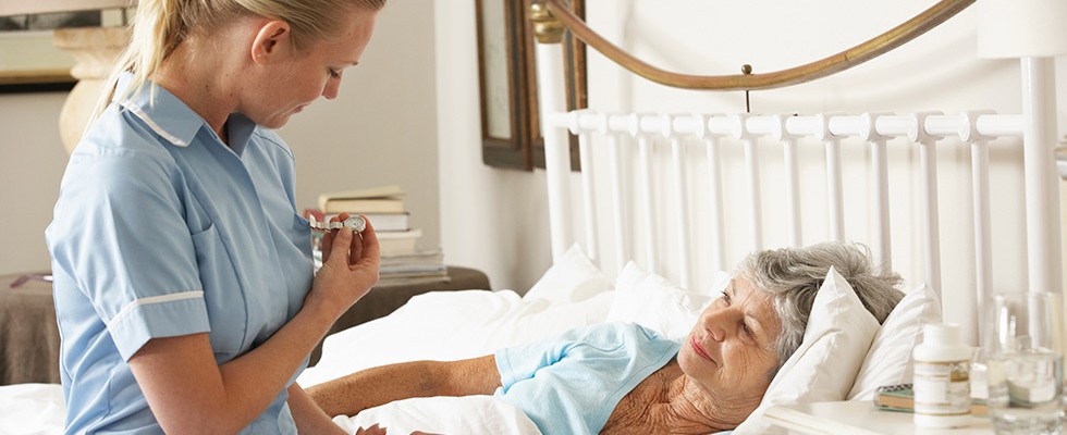 An older woman lies down in bed while a caregiver checks her heart beat.