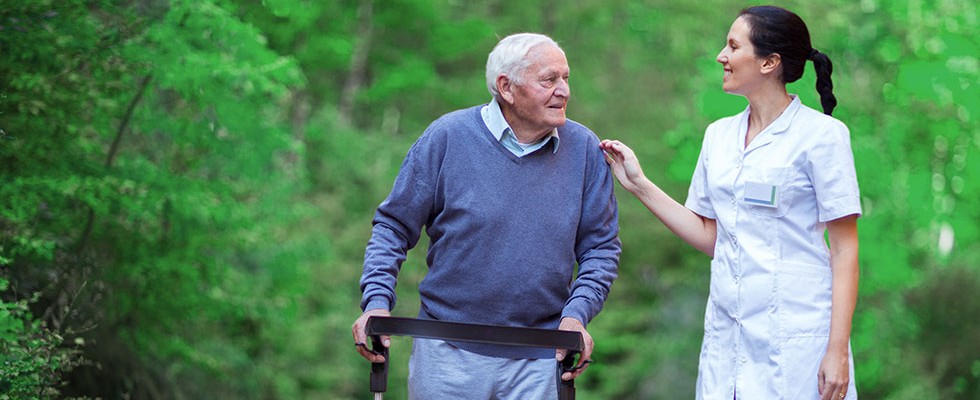 An older man uses a rollator to assist his mobility while he goes on a walk with his caregiver outside. 