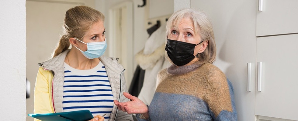 A blonde woman with a mask and a clipboard looks at an older woman with a mask who appears to be gesturing dramatically and looking upward.