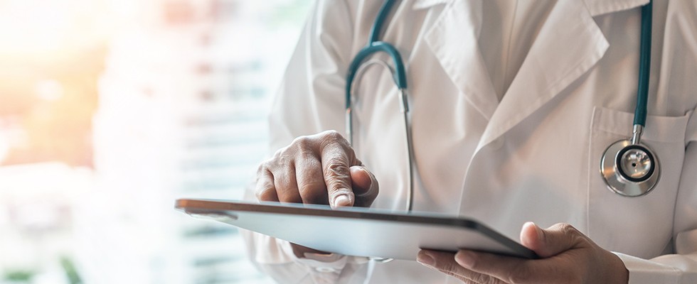 Image of person in white lab coat with stethoscope around their neck looking at and touching an ipad. 