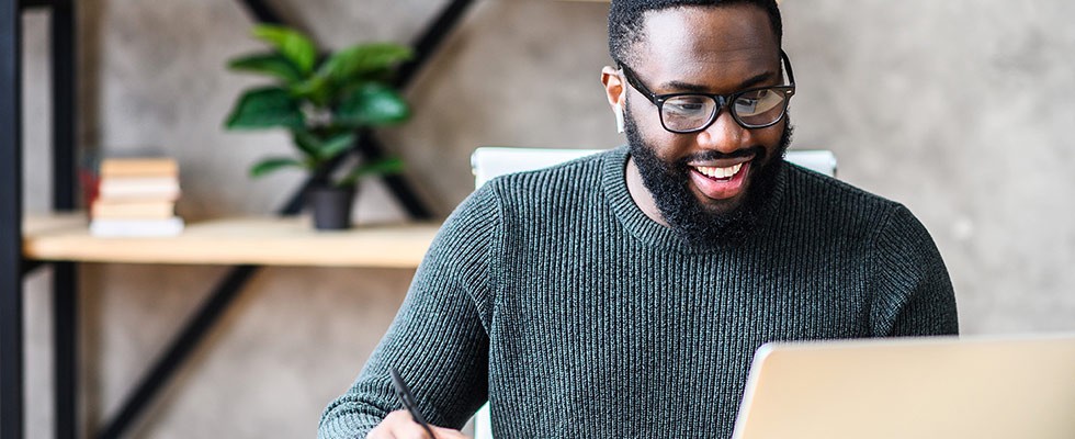 A smiling man with glasses is working with air pods in in front of a laptop while writing something down. 