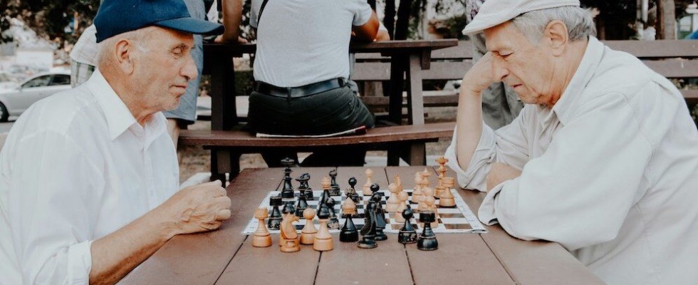 Two elderly men in white shirts playing a game of chess outside on a wooden table.