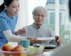 A young woman caregiver is bringing a tray of breakfast food to an older gentleman in his home.