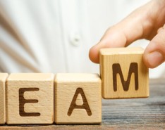 A man sits behind a wooden table with three wooden blocks in front of him with the letters T, E, and A on them. He is setting down another with the Letter M, to spell out the word "Team".