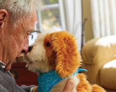 An older man holds a companion pet, which looks like a brown and white spotted dog with a blue bandanna around its neck, up to his face. Touching his nose with the robotic dogs. 