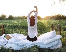 A young woman stretches while sitting on her bed in the middle of an open field.