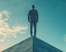 An image of a man in a suit from the back, who is standing on top of a pyramid. A blue sky with clouds is in the background. 