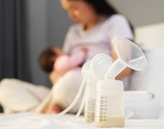 In the foreground, a breast pump is sitting with milk in it. In the background a woman is nursing a baby..