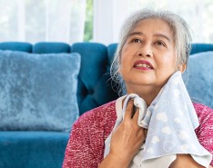 An older woman sits in front of a couch, with a cloth to wipe away sweat.