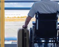 Wheelchair user at an airport with suitcase, looking out a window at the runway.