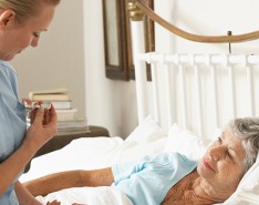 An older woman lies down in bed while a caregiver checks her heart beat.