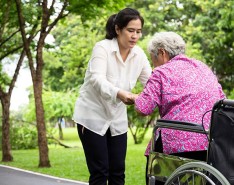 An older in woman in pink is being assisted to stand up from her wheelchair by a woman in white. The two are outside. 