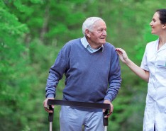 An older man uses a rollator to assist his mobility while he goes on a walk with his caregiver outside. 