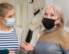 A blonde woman with a mask and a clipboard looks at an older woman with a mask who appears to be gesturing dramatically and looking upward.