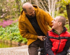 A man in yellow assists another man in red who is in a wheelchair. The two are smiling at each other and outside. 