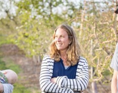 A family (two men and a woman) stand in a field. One of the men holds a baby.