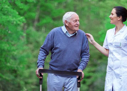 An older man uses a rollator to assist his mobility while he goes on a walk with his caregiver outside. 